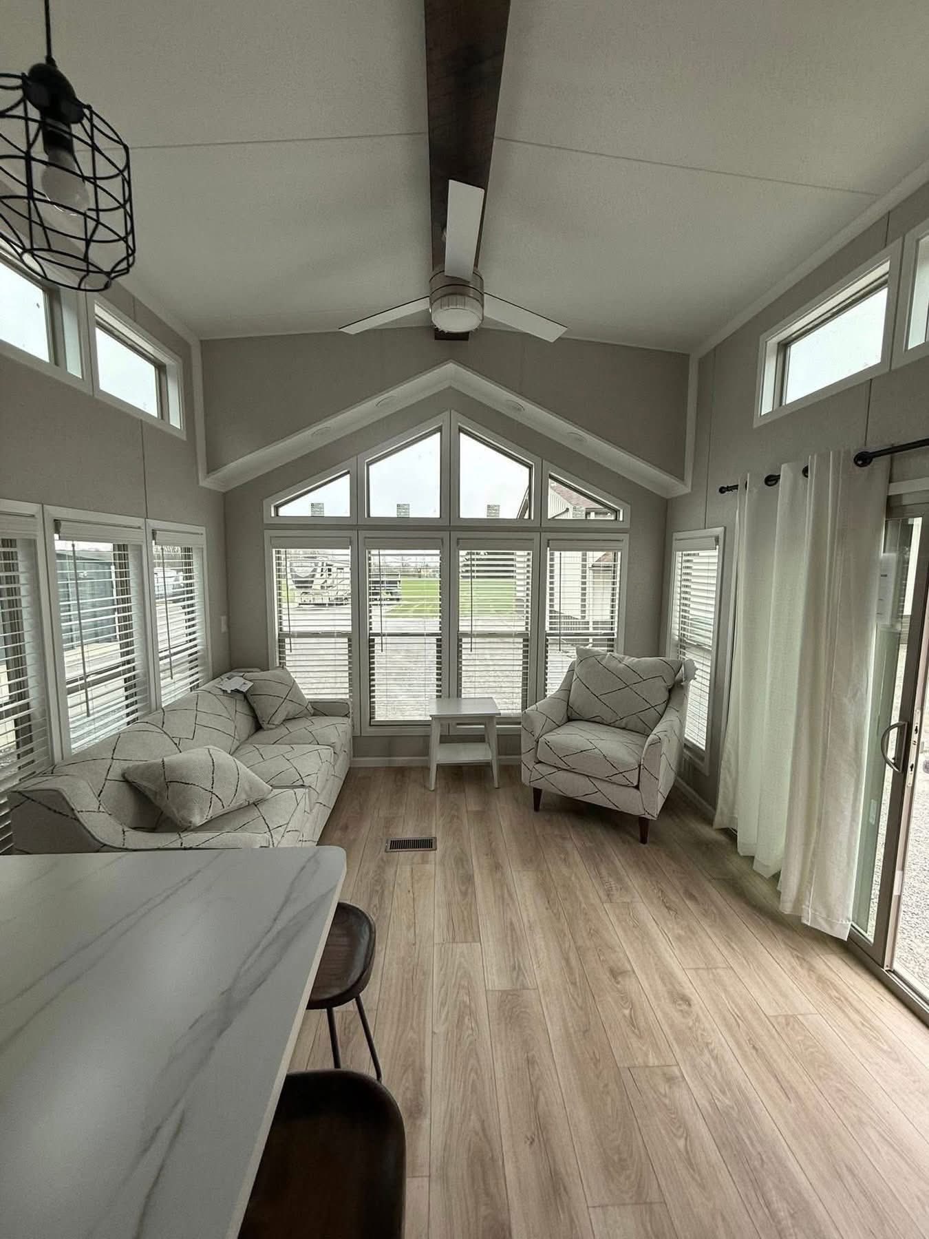Bright, modern living room with large windows, a triangular ceiling, and light wood floors. Features patterned sofas, a chair, and neutral-toned decor.