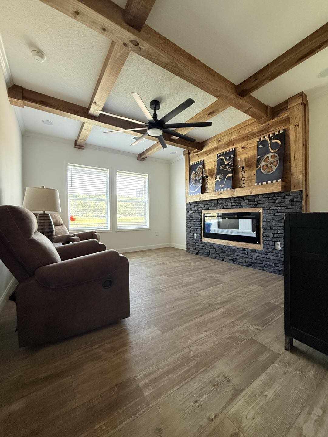 Living room with a rustic vibe, featuring brown recliners, a wooden beamed ceiling, light wood floors, and a stone fireplace with art above, creating a cozy feel.