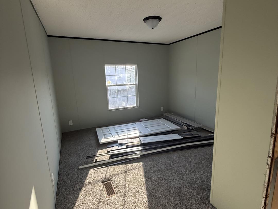 Empty room with light gray walls and dark carpeting. A single window lets in sunlight. A white door and metal frames are stacked on the floor.