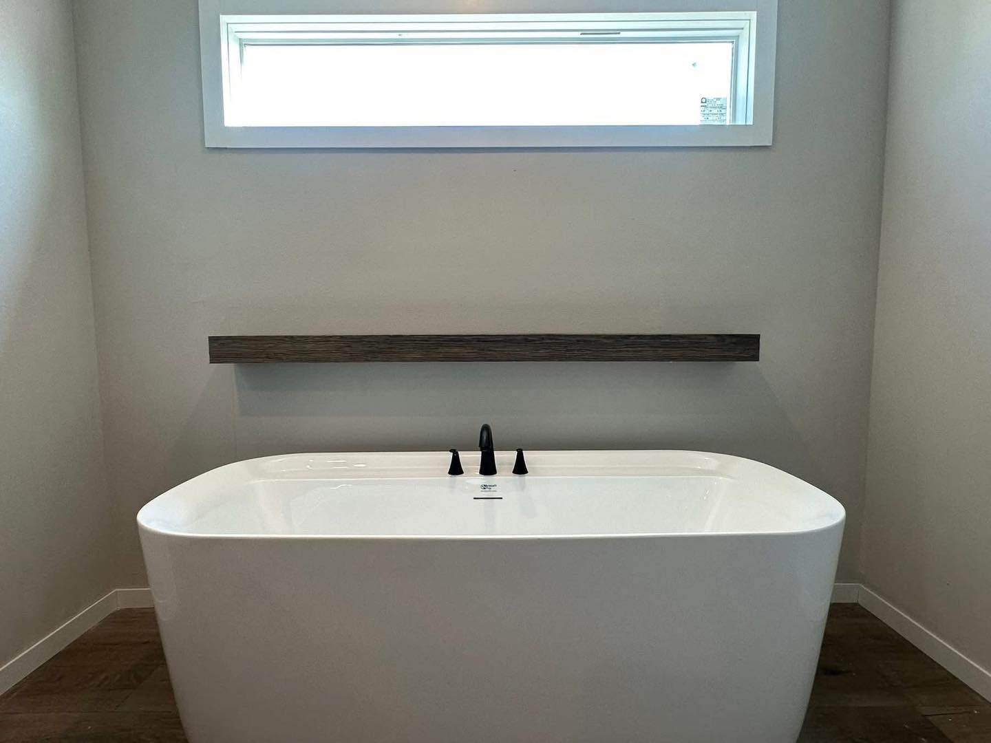 Minimalist bathroom with a white freestanding bathtub, black fixtures, and a wooden shelf above. Soft natural light from a narrow window creates a serene atmosphere.