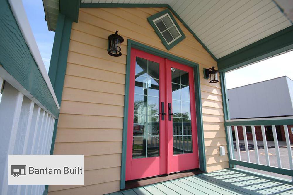A cozy porch with a vibrant pink double door, yellow siding, and teal trim. Black lanterns add charm, evoking a welcoming and cheerful vibe.