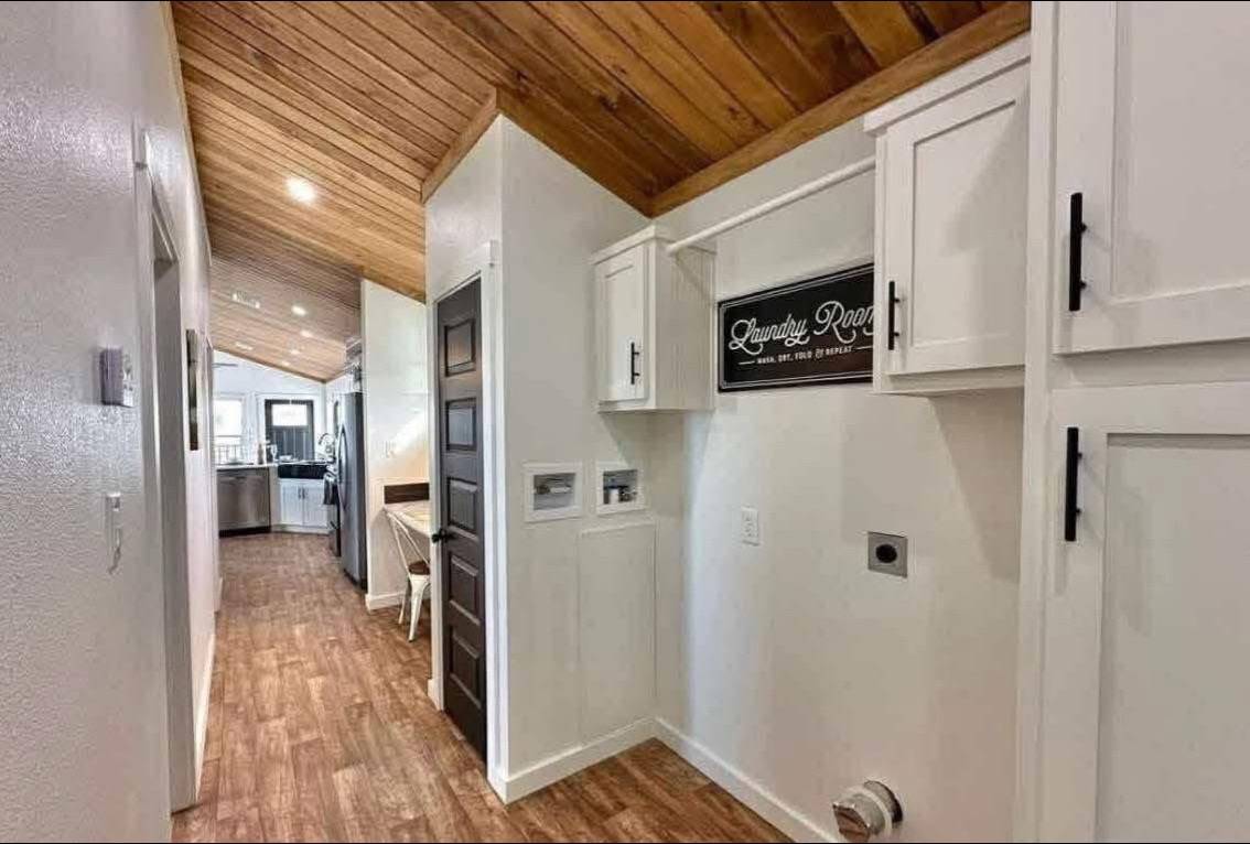 Hallway with wooden ceiling and floor, leading to a kitchen. White cabinets and a “Laundry Room” sign hang on the wall, creating a cozy, rustic vibe.