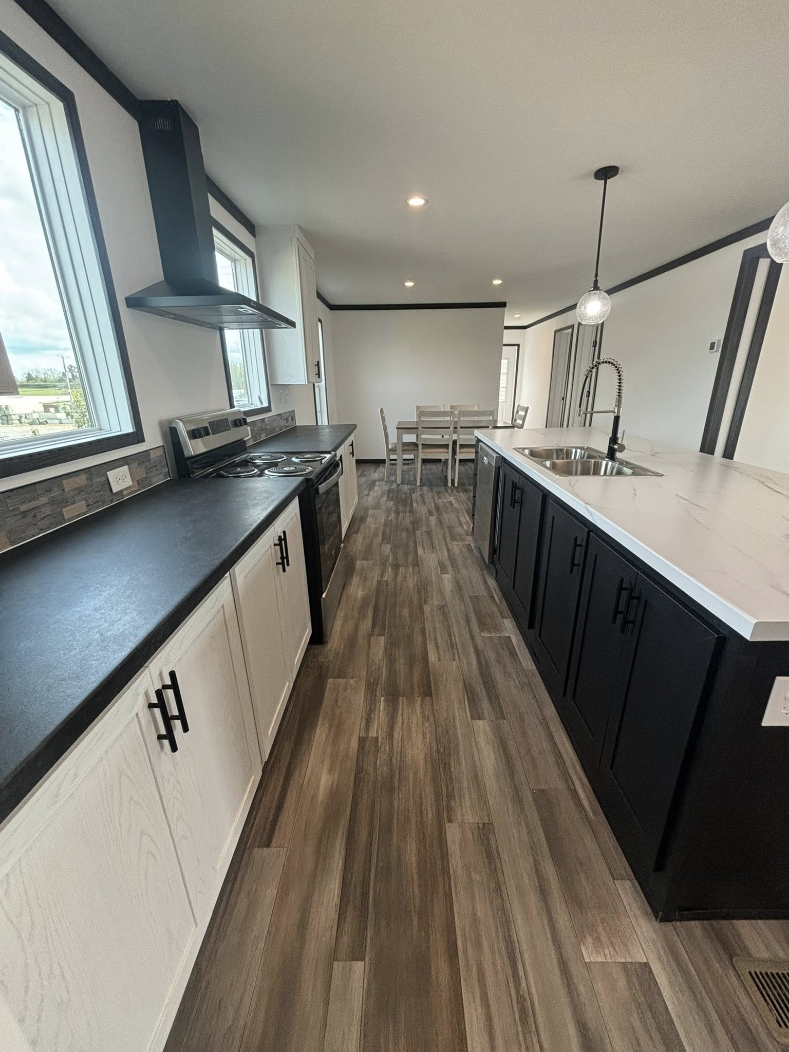 Modern kitchen with sleek black and white design, featuring a central island with a sink, wood flooring, and a dining table in the background. Bright and spacious.