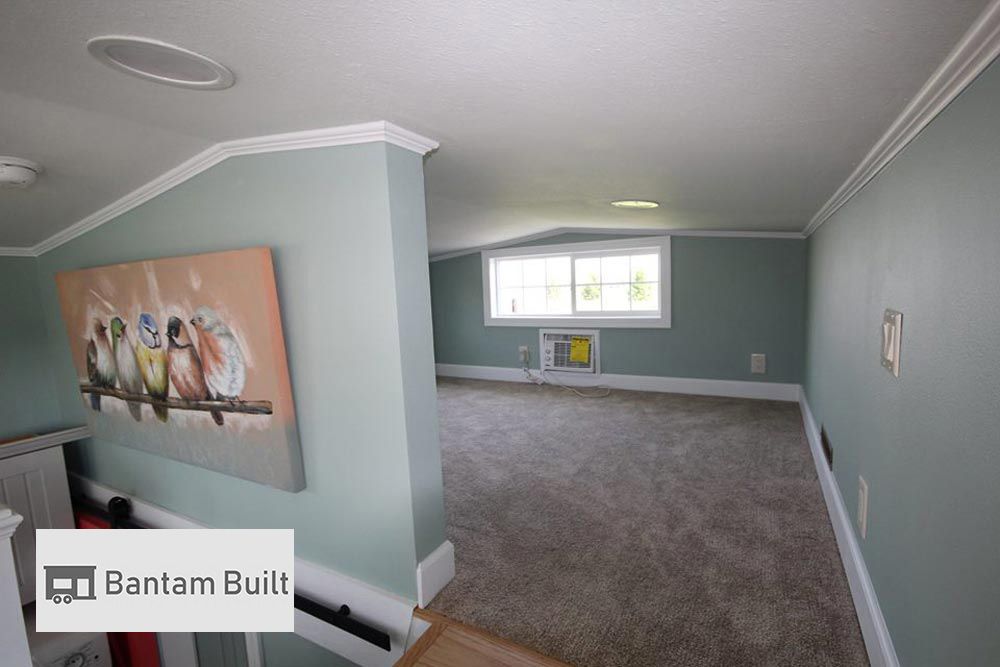 Small loft space with light green walls and carpeted floor, featuring a painting of colorful birds. A window lets in natural light, creating a cozy ambiance.