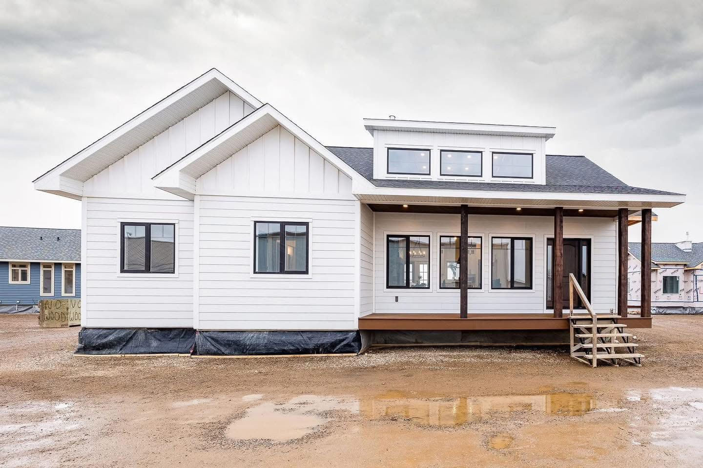 A white modern house with a gabled roof and dark trim sits on a muddy plot. The front porch has wooden posts and steps, under a cloudy sky.