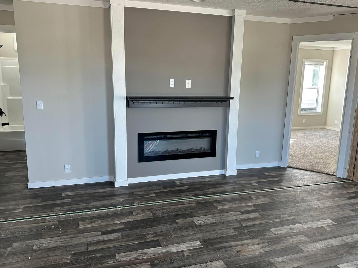 Modern living room with gray walls and dark wood flooring. Features a sleek electric fireplace, white trim, and an adjacent carpeted room with a large window.