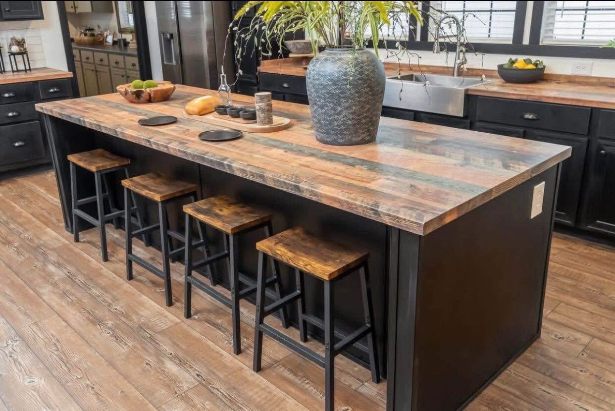 A modern kitchen with a large wooden island features five high stools. A blue vase with plants and a bowl of fruit add a fresh touch. Warm and inviting tone.