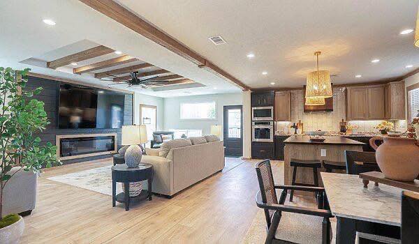 Spacious open-plan living room and kitchen with wooden beams, modern furnishings, and warm lighting. A large TV, cozy sofas, and dining table add comfort.