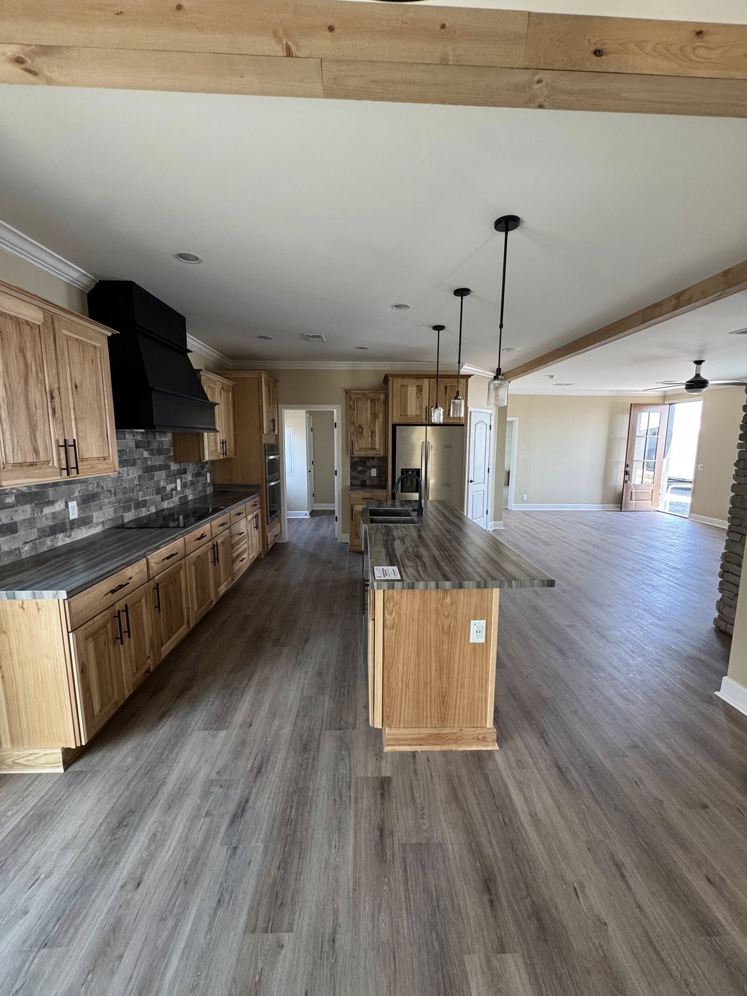 A spacious kitchen features wooden cabinets, a stone-textured backsplash, and a central island under pendant lights. Adjoining is an open living area with wooden floors.