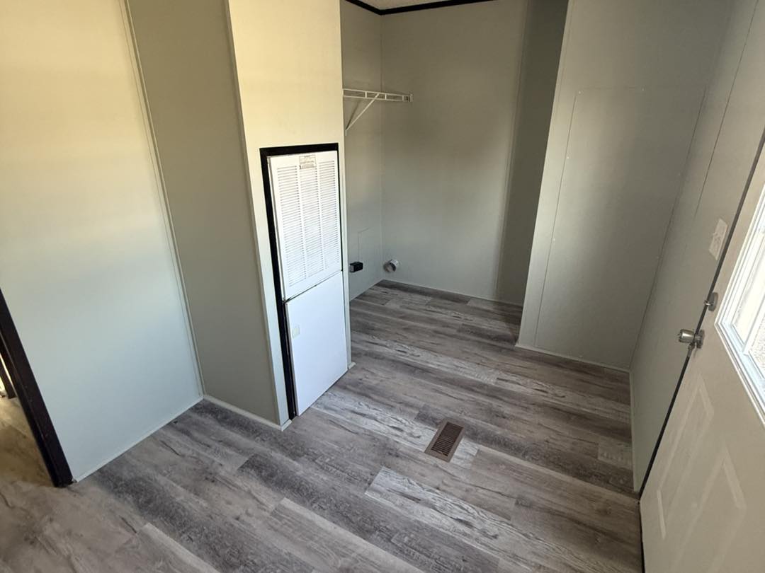 Simple, empty room with gray walls and wood-patterned vinyl flooring. Bright light enters through a window near a white door, creating a neutral atmosphere.