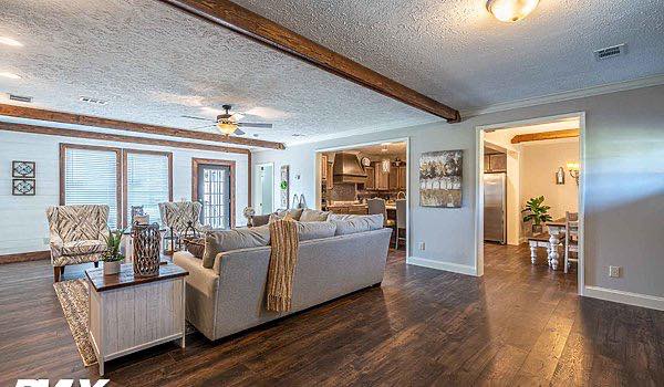 Spacious living room with hardwood floors and wooden beams. Cozy gray couches, patterned armchairs, and an open kitchen create a warm, inviting space.
