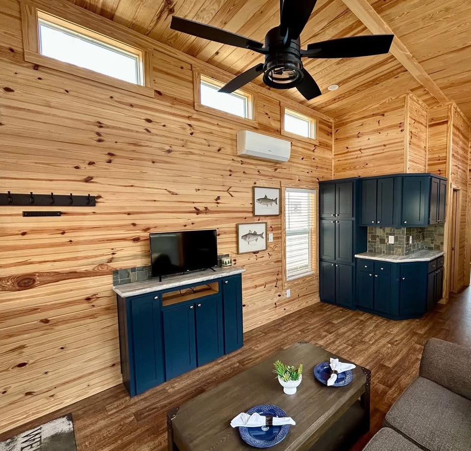 Cozy wooden interior with high ceilings, featuring a ceiling fan, small TV on a blue console, and blue kitchen cabinets. The ambiance is warm and inviting.