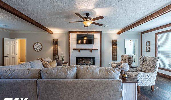 Modern living room with a beige sectional sofa facing a fireplace and wall-mounted TV. Features exposed wooden beams, a ceiling fan, and patterned armchairs, creating a cozy ambiance.