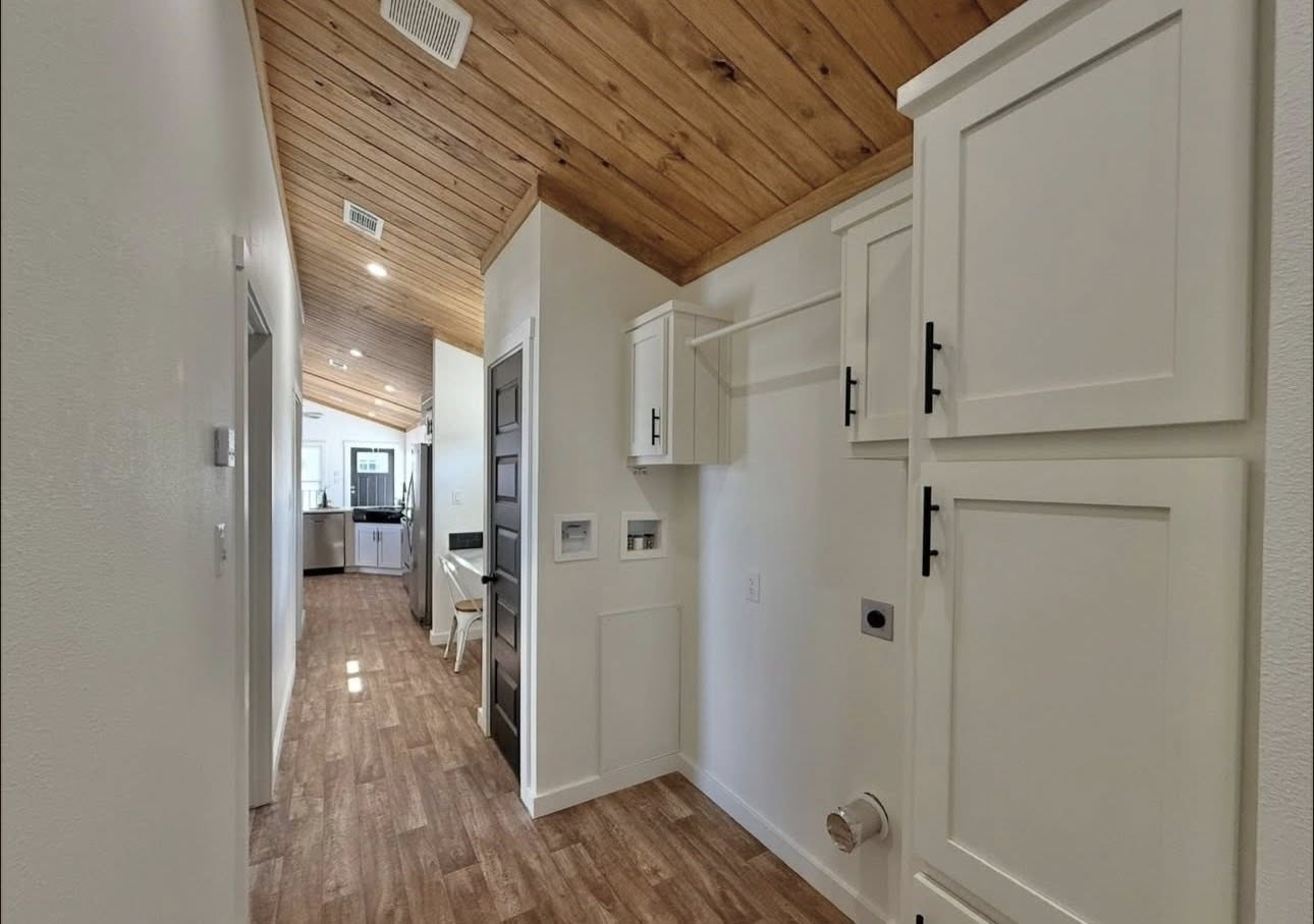 Narrow hallway with wooden ceiling and floor, white walls, and cabinets to the right. Leads to a bright kitchen with stools, conveying a cozy, modern feel.
