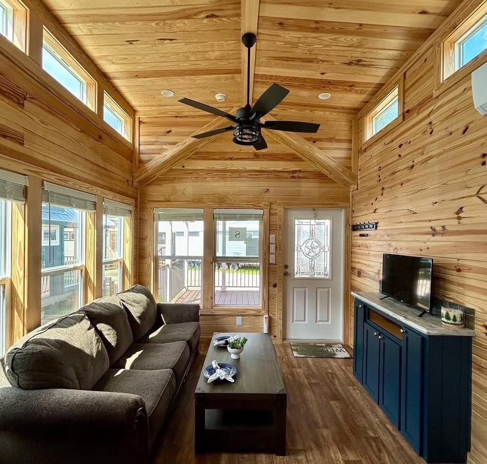 Warm, cozy living room with wooden walls and ceiling, large windows, gray sofa, dark coffee table, ceiling fan, TV on blue cabinet, and glass door.