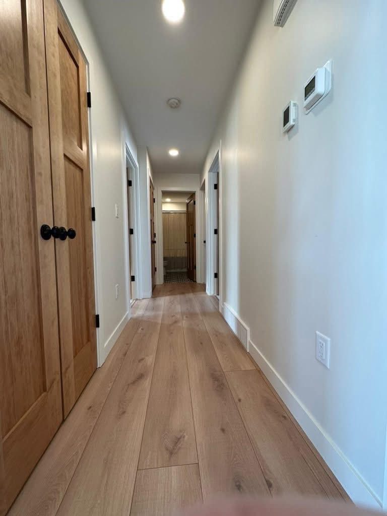 A long, clean hallway with light wood flooring and white walls. Wooden doors are on both sides, and a bathroom is visible at the far end. Bright lighting.