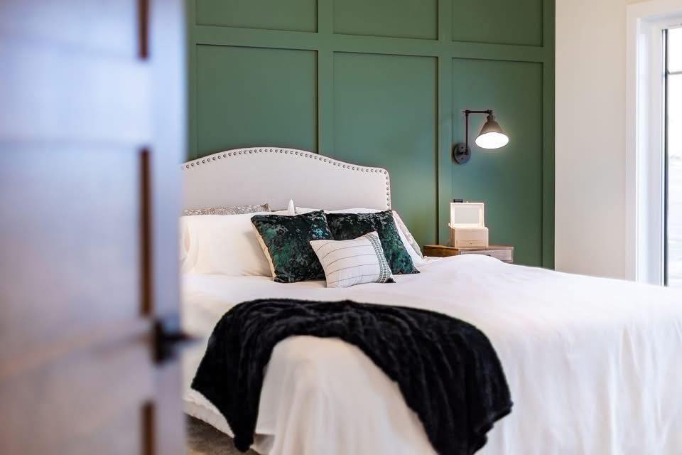 Cozy bedroom with a white bed, green accent pillows, and a dark blanket. A green paneled wall and a sconce add elegance and warmth.