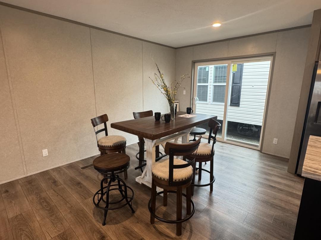 A cozy dining area features a wooden table with four cushioned chairs, centered under warm lighting. A vase with branches adds elegance. Glass doors reveal an exterior view, enhancing the room's welcoming ambiance.