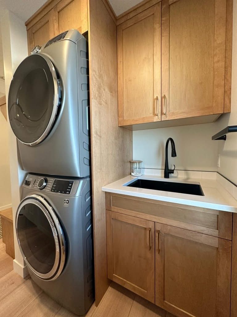 Compact laundry room with stacked silver washer and dryer adjacent to a light wood cabinet and counter with a black faucet and sink. Cozy and efficient space.