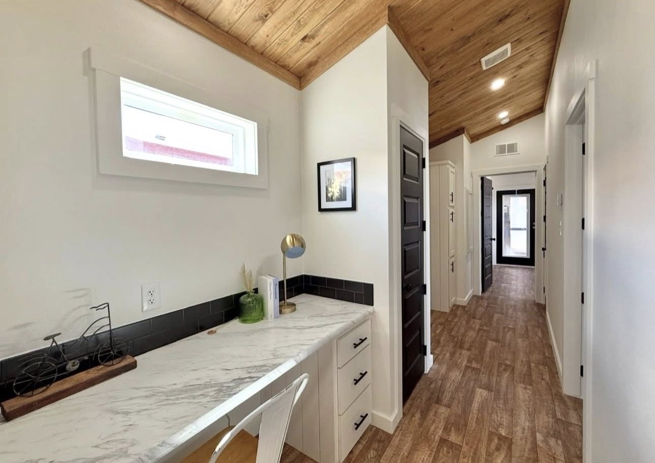 A cozy hallway with light wood flooring and ceiling, featuring a marble desk with a chair. A small window and framed picture add charm. Modern and inviting.