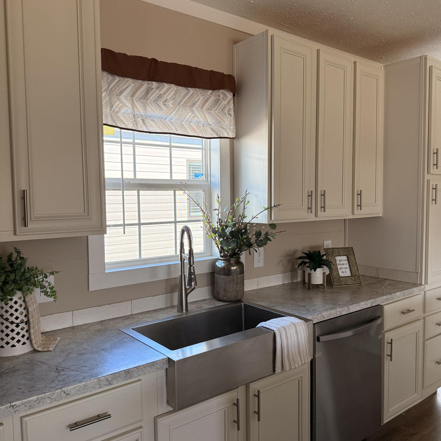 Bright kitchen with light cream cabinets and a stainless steel farm sink under a window. Decor includes potted plants, a framed picture, and a vase.
