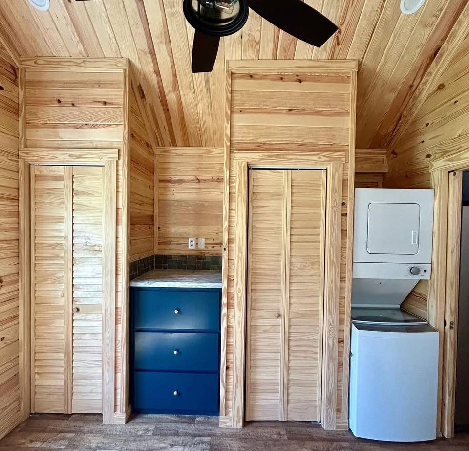 Compact wooden room featuring vertical wood paneling, a stacked washer-dryer, a set of blue drawers, and a ceiling fan, conveying a cozy cabin feel.