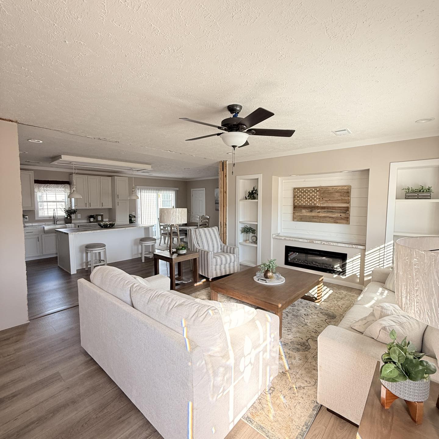 Spacious, bright living room with light-colored sofas, a wooden coffee table, and potted plants. A ceiling fan hangs above; the adjoining kitchen is visible.