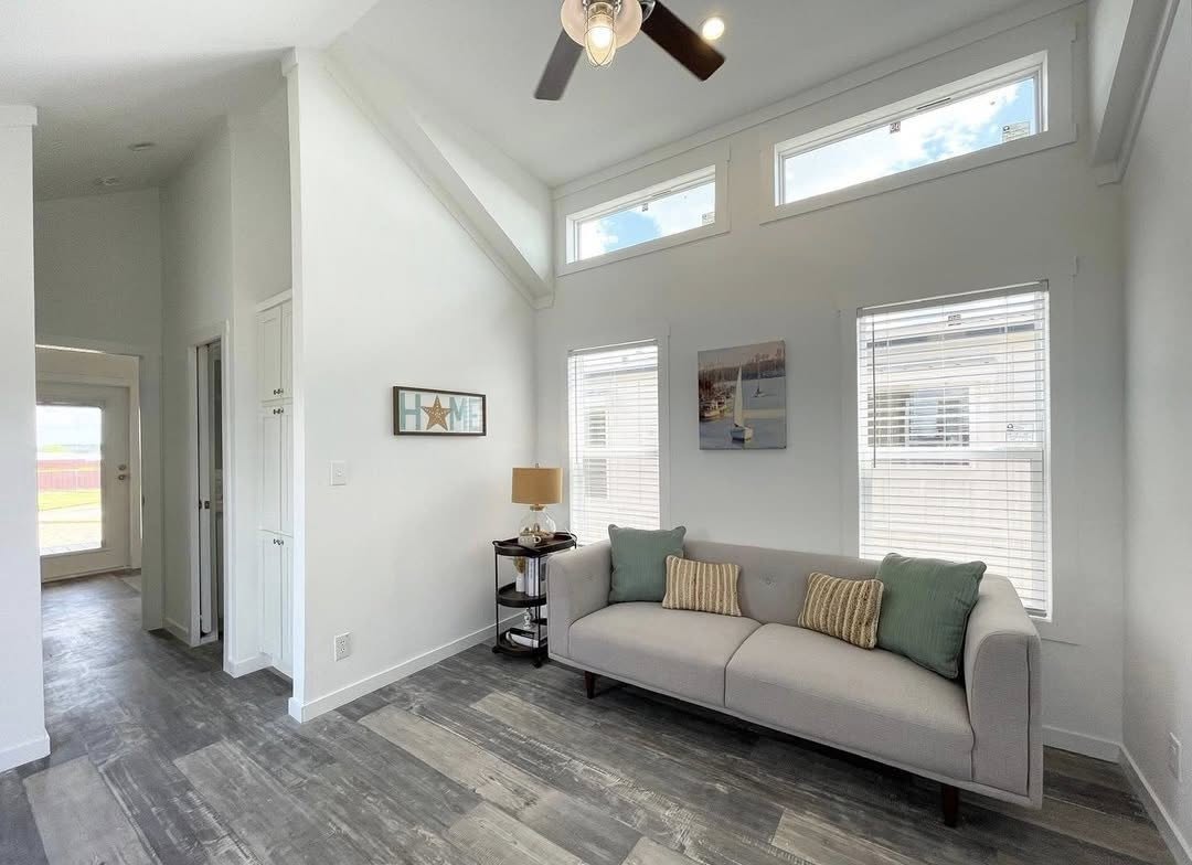 Bright living room with a sleek gray couch adorned with patterned cushions. Sunlight streams through tall windows, revealing light wood flooring and a fan above, creating a cozy and airy atmosphere.