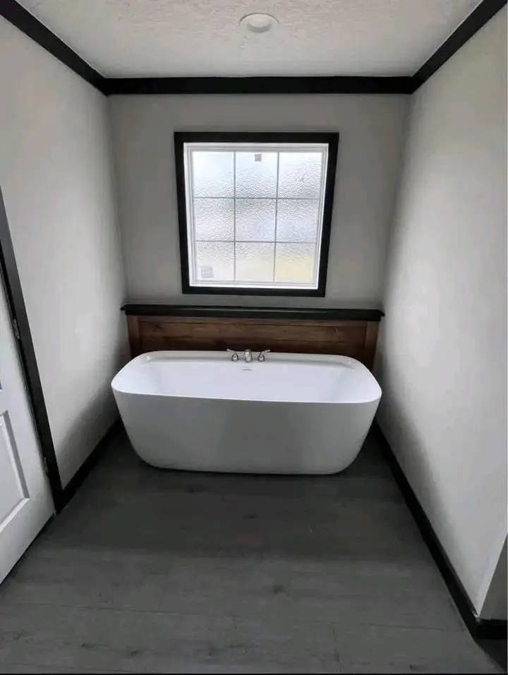 Minimalist bathroom with a white freestanding bathtub centered under a frosted window. The room has light gray walls and dark flooring, creating a calm atmosphere.