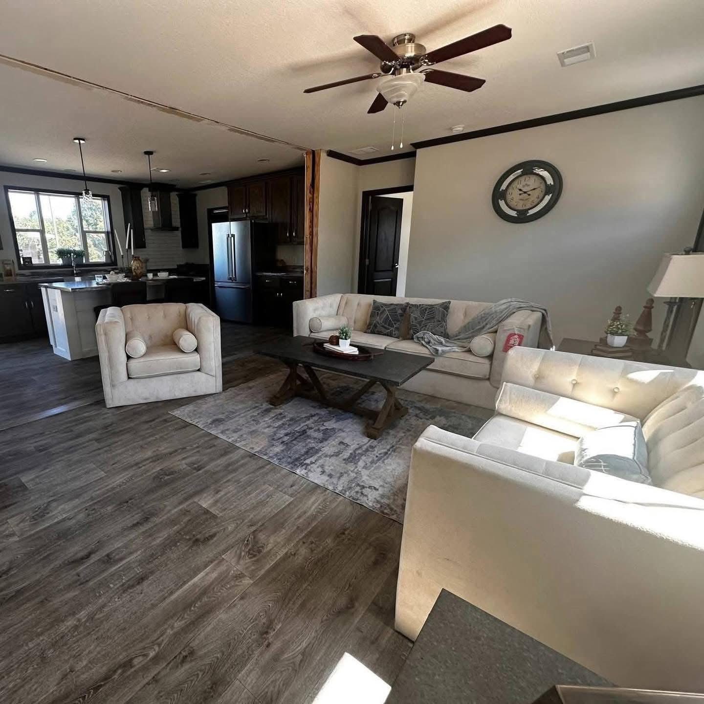 Stylish living room with beige sofas around a wooden table, gray rug, and ceiling fan. Open kitchen with dark cabinets visible. Cozy and welcoming.