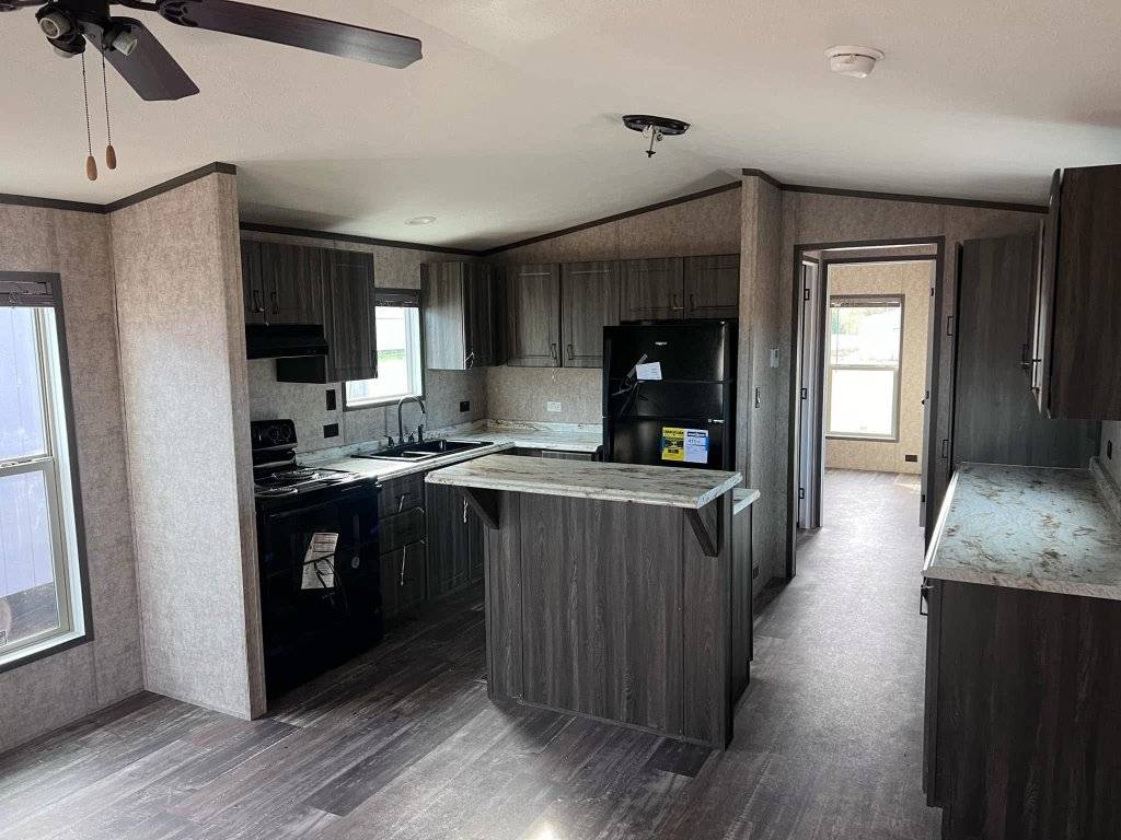 Modern kitchen with dark wood cabinets, black appliances, and a small center island. Bright natural light filters through large windows, creating a cozy atmosphere.