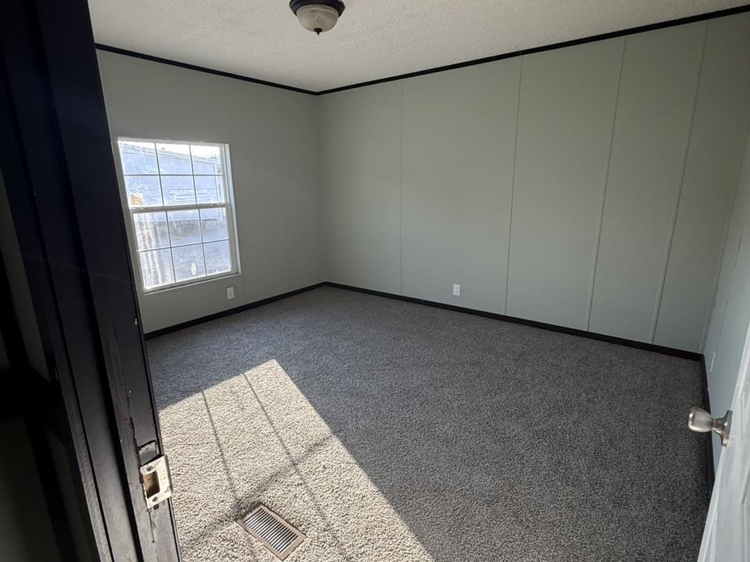 Empty room with gray walls, a carpeted floor, and a single window casting sunlight inside. The atmosphere is calm and minimalistic.