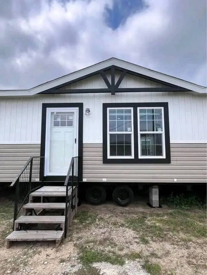 Compact modular home with beige siding, black trim, and a small front porch. The home has a white door and double windows. Overcast sky above.