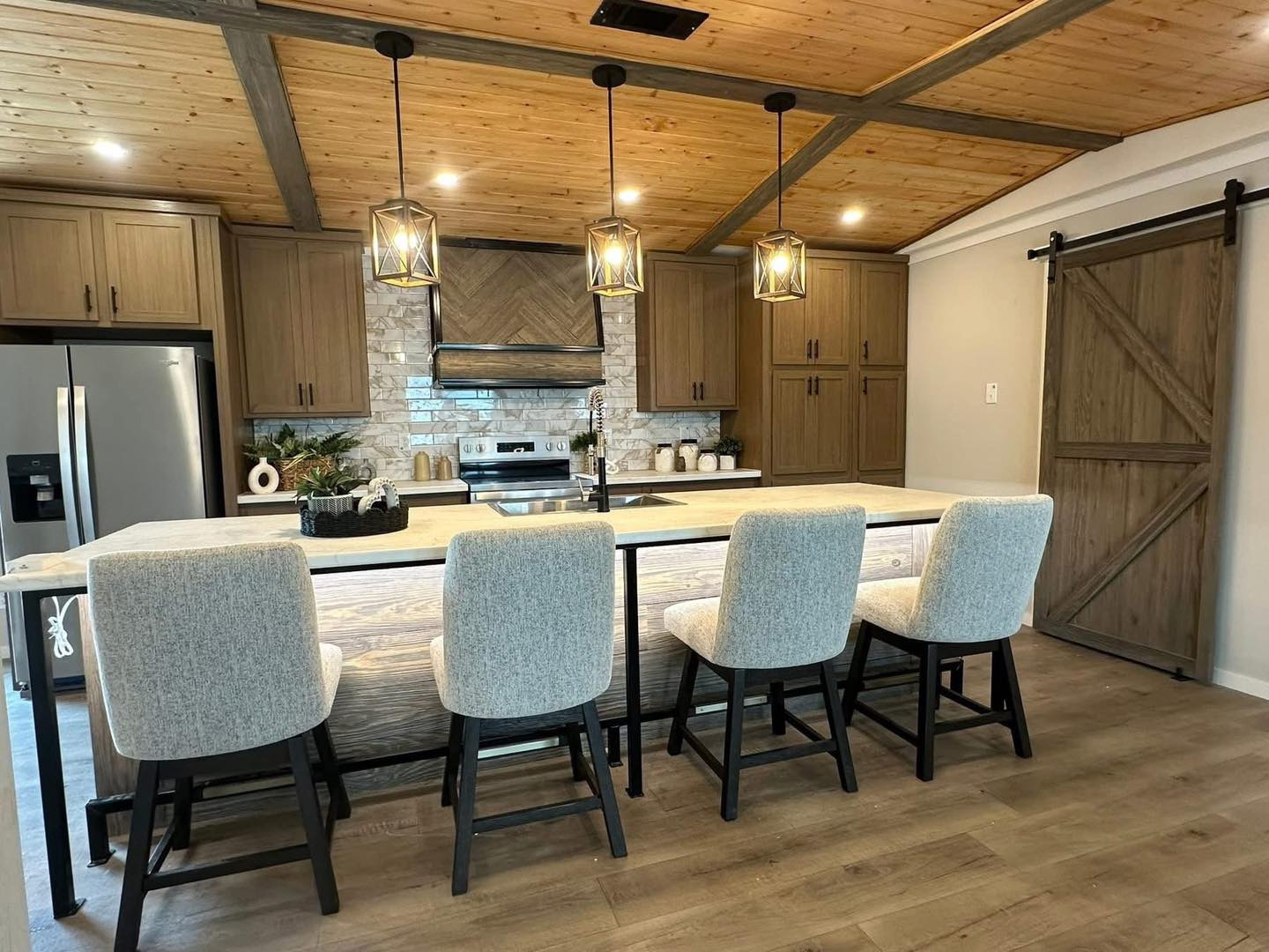 Modern kitchen with light wood cabinetry, stone backsplash, and a central island. Four cushioned stools, pendant lights, and a sliding barn door create a cozy, rustic ambiance.