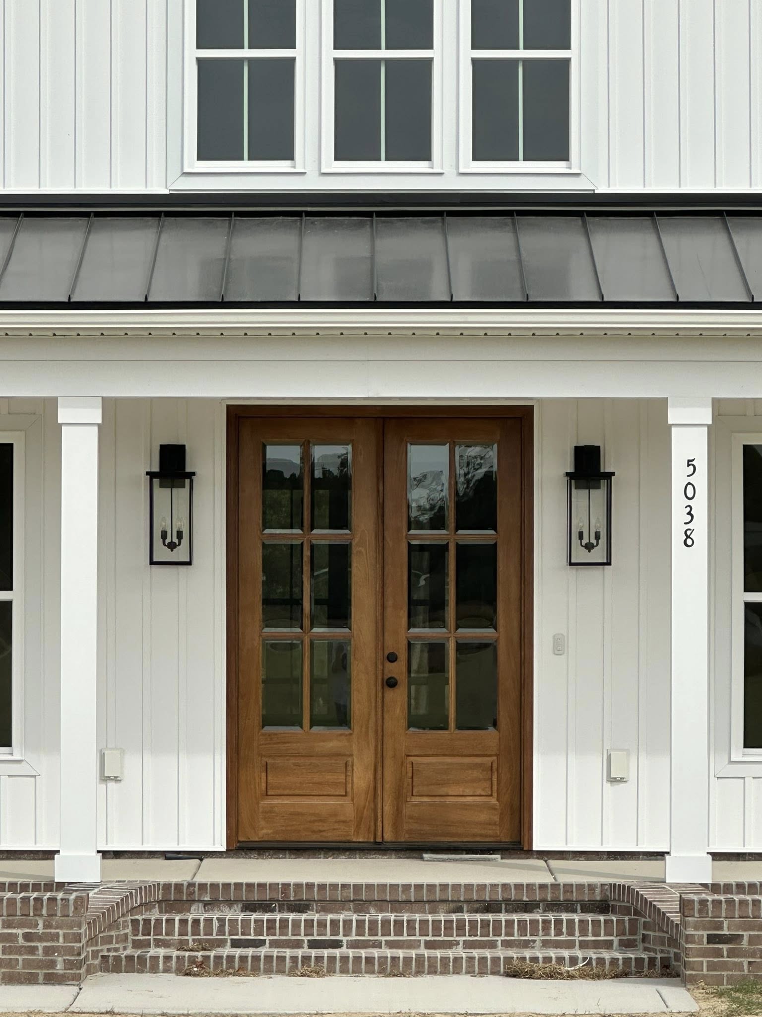White house exterior with a large wooden double door flanked by tall glass lanterns. Above, a row of windows. Brick steps lead to the entrance.