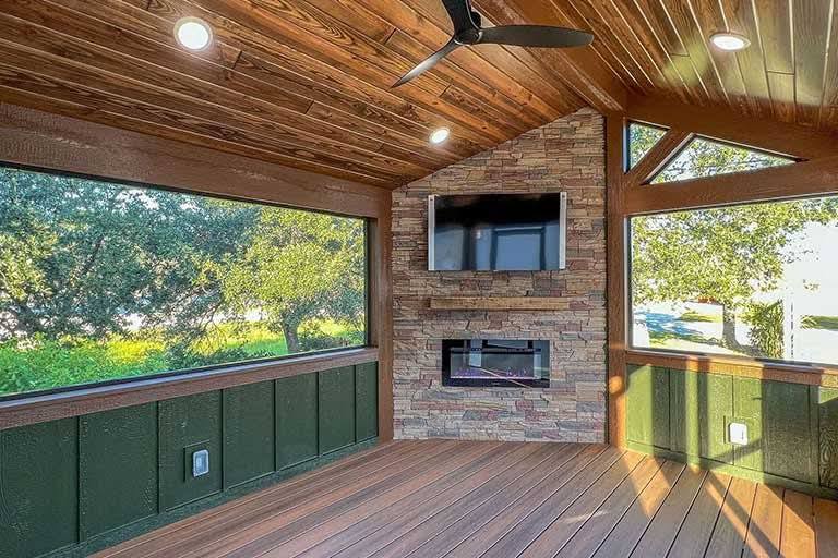 Cozy sunroom with wooden floors and ceiling, featuring a stone wall with an electric fireplace and TV. Large windows showcase lush greenery outside.