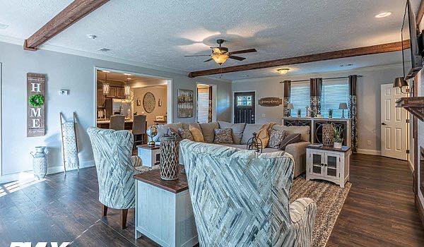 A cozy living room with a rustic charm, featuring a gray sofa, patterned armchairs, and wooden beams on the ceiling. The space is warmly lit, with large windows allowing natural light. The open layout leads to a kitchen visible in the background. A decorative "HOME" sign hangs on the wall, adding a welcoming touch.