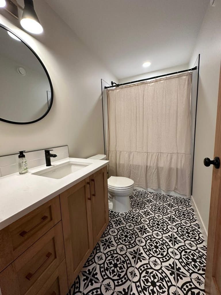 A modern bathroom with a wooden vanity and white countertop, black faucet, round mirror, patterned black and white tile floor, and beige shower curtain.