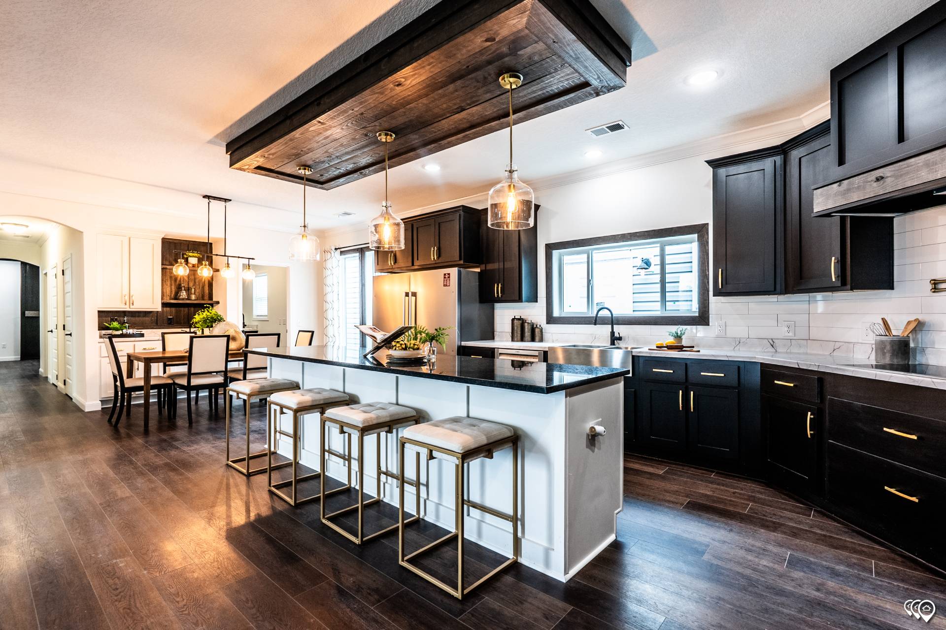 Modern kitchen with dark wood cabinets, a large island with four stools, and pendant lights. Dining area visible on the left. Bright and inviting space.