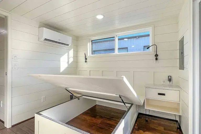 A bright, minimalistic room with white paneled walls and wood flooring. An open storage bed reveals its wooden interior. A small table and window are present.