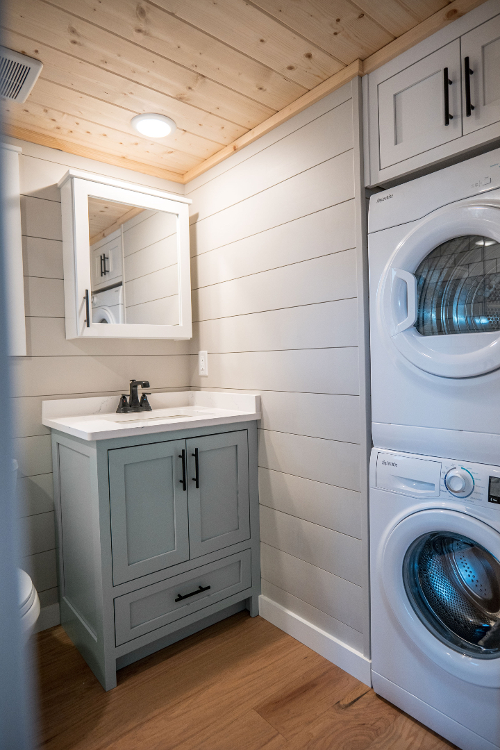 Compact laundry room with a wooden ceiling and light gray shiplap walls. Features a small vanity with a mirror, and a stacked washer and dryer.