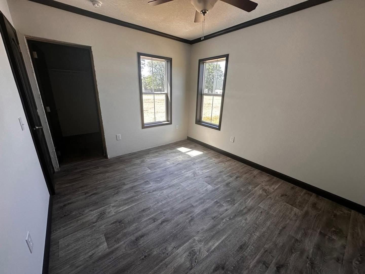 A small, empty room with gray laminate flooring, beige walls, and two windows letting in natural light. There is a ceiling fan and an open door to a closet.