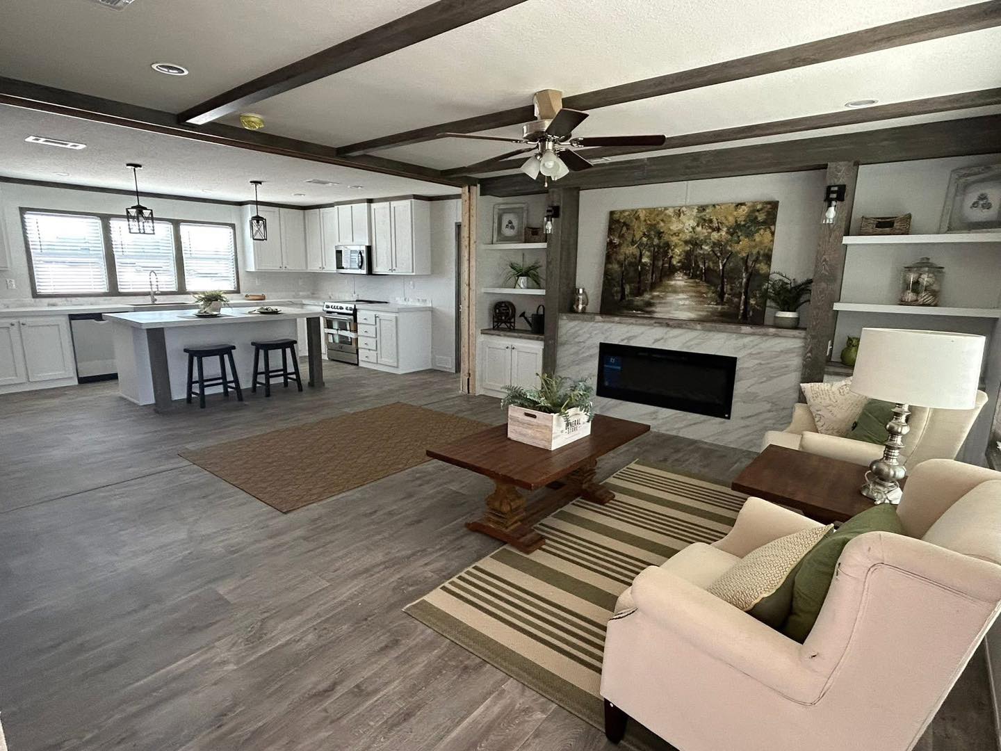 Spacious living area with modern kitchen, featuring a central island and bar stools. Cozy seating area with a striped rug, armchair, and fireplace art.