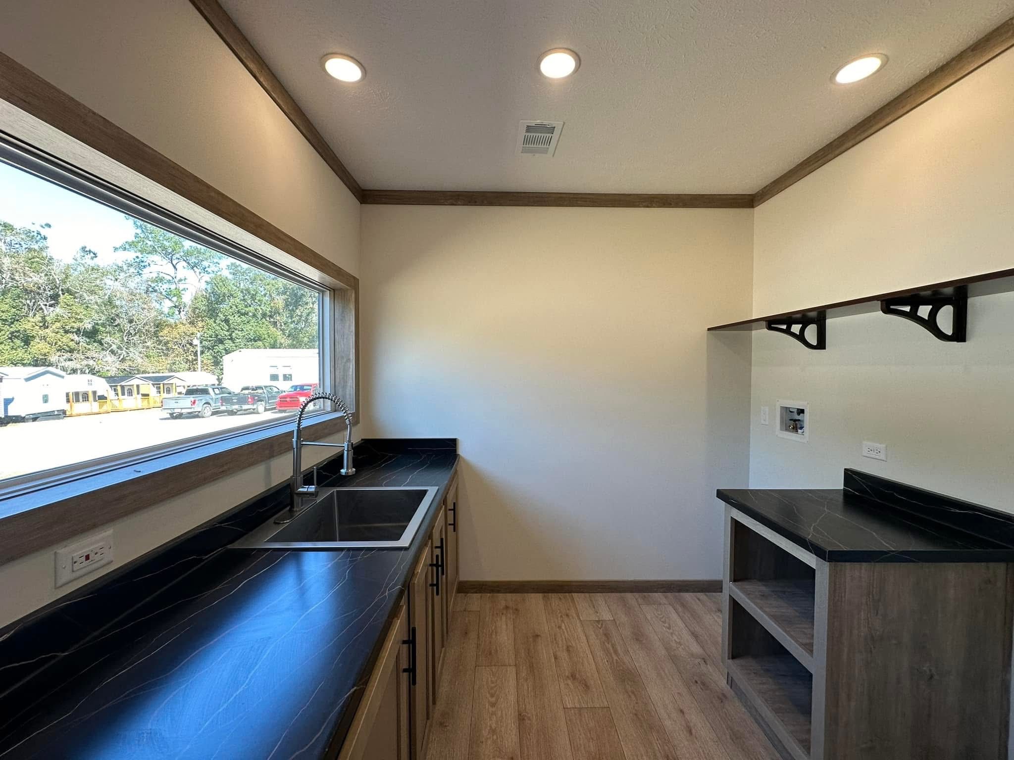 Modern kitchenette with black countertops, light wood floors, and upper open shelves. Large window view of parked cars and trees. Clean, minimal design.