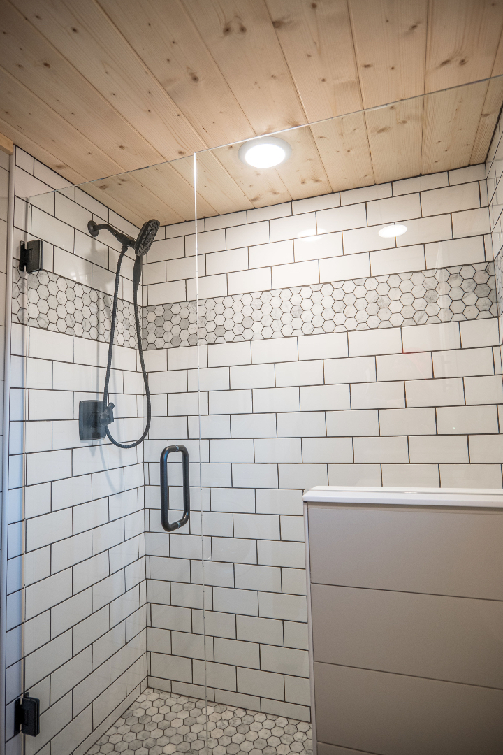 Modern shower with white subway tiles and a hexagonal mosaic strip. Features a sleek black showerhead, glass door, and wood ceiling, creating a fresh, clean ambiance.