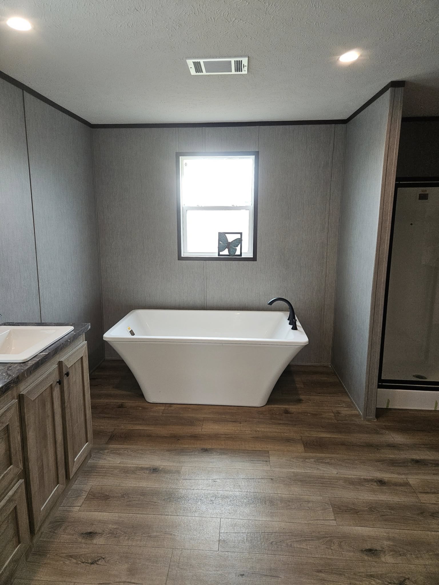 Modern bathroom with a freestanding white tub under a window, gray walls, wood-look flooring, and a sleek black faucet. Calm and minimalist design.