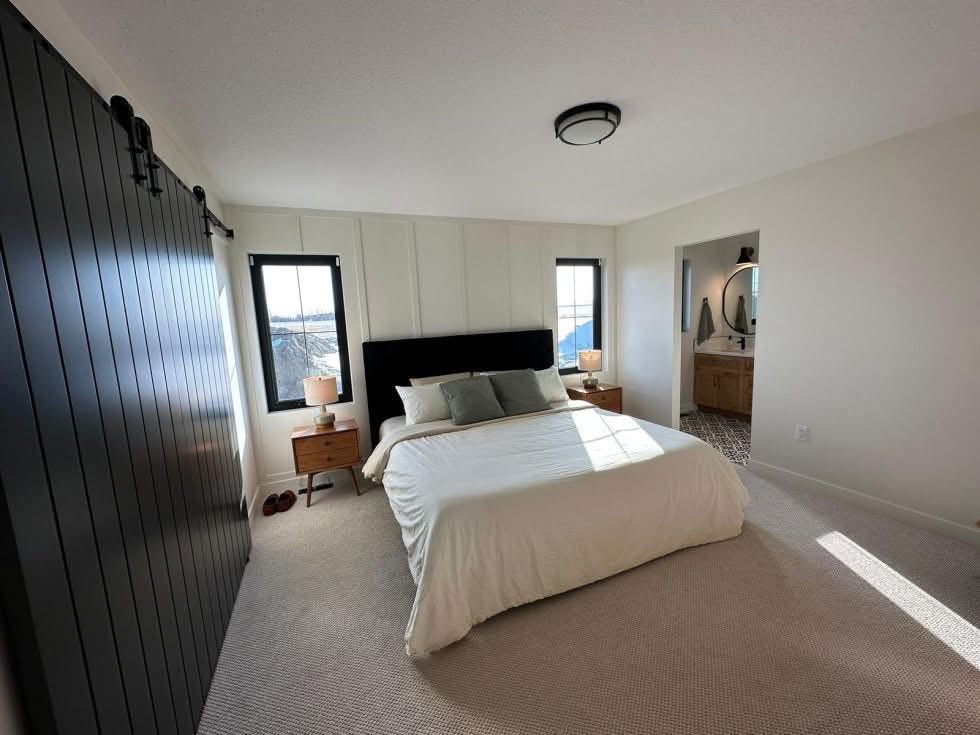 Modern bedroom with a white bedspread, gray pillows, and wooden nightstands beside a black headboard. Sunlight streams through two windows, creating a bright and airy atmosphere. A dark accent wall contrasts with light walls and carpet. An open doorway shows a glimpse of a stylish bathroom.