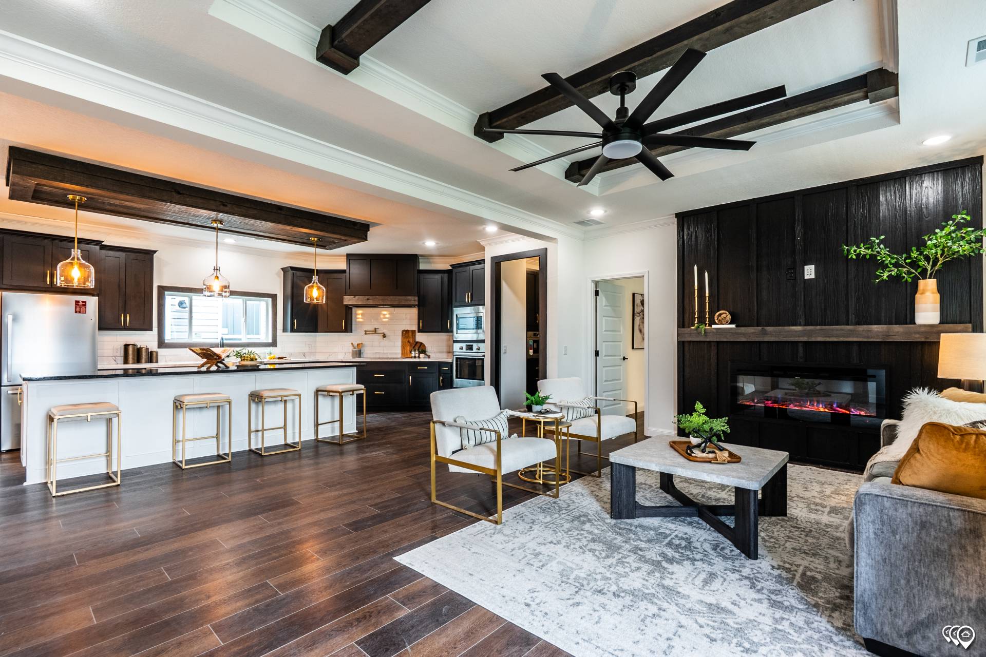 Stylish open concept living room and kitchen with warm wood floors, dark cabinets, and modern furniture. Features a large fan, cozy fireplace, and ambient lighting.