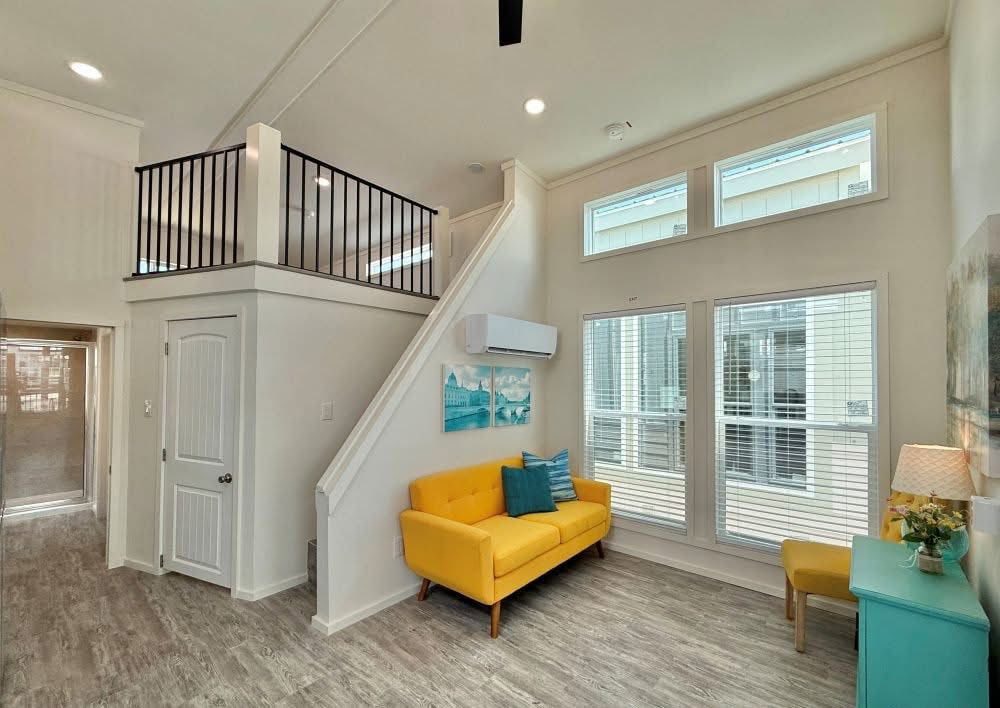 Bright living room with high ceilings, featuring a vibrant yellow sofa, teal pillows, modern artwork, and large windows providing natural light. Cozy, welcoming feel.