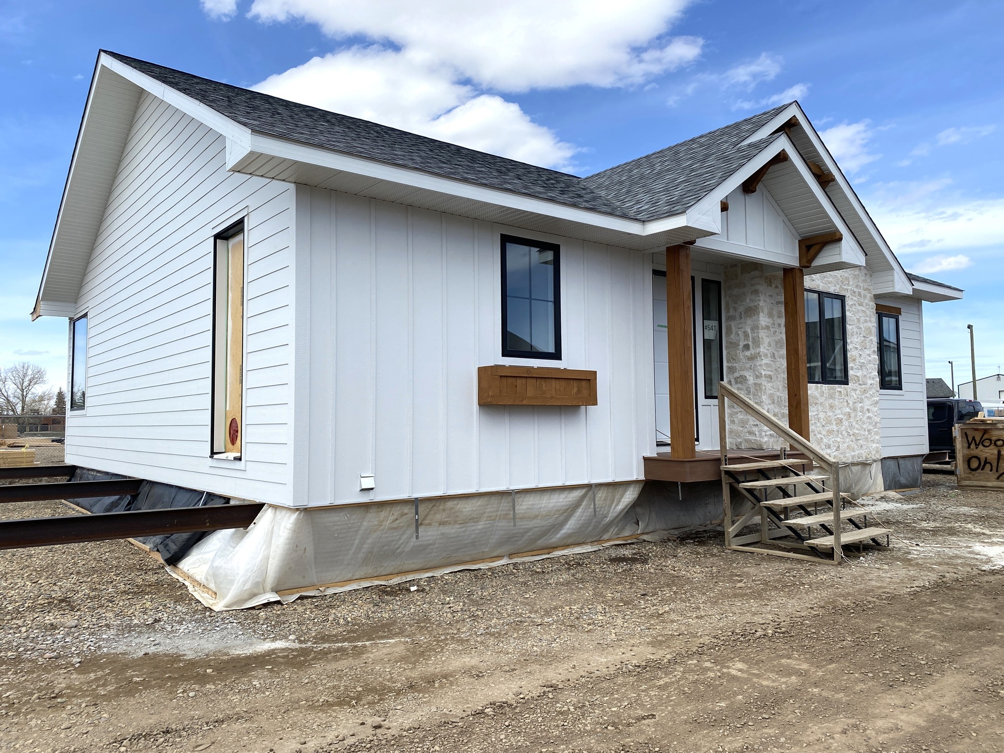 Single-story, modern white house with stone accents and a gray roof under a blue sky. Front steps lead to a central door. Construction materials around.